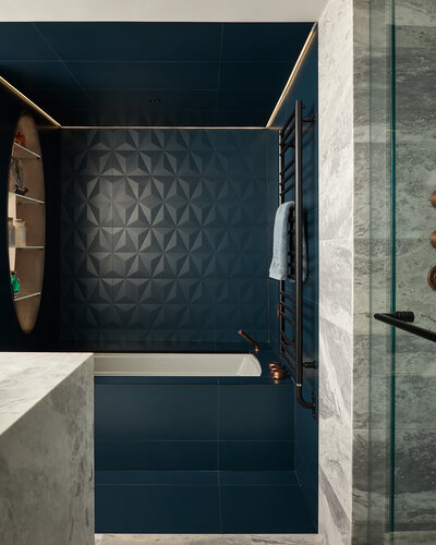 A luxury bathroom featuring built-in cove lighting, a tub surrounded by deep blue 3D tiles, and a marble countertop vanity and shower area