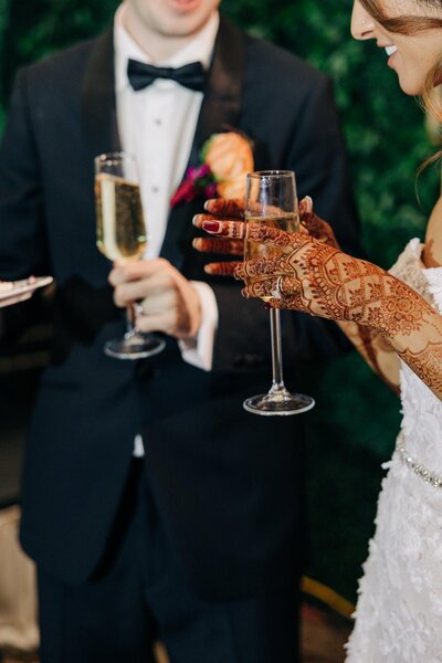A bride and groom with a glass of champagne, the bride's hands adorned with henna tattoos.