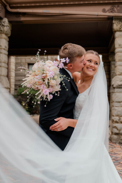 Groom kisses bride on the check while bride smiles past the camera with her veil flowing in the wind.