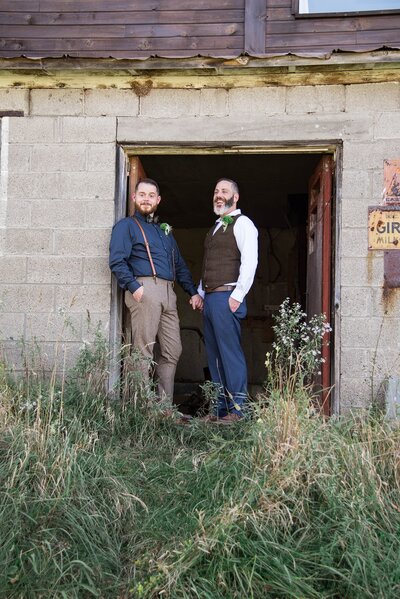 Two grooms standing together in a rustic doorway.
