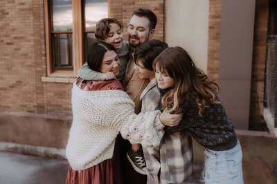 A group of five people, including two adults and three children, stand closely together outside near a brick building, smiling and hugging each other.