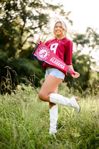 cuthbertson high school grad holding university alabama pennent and wearing a jersey, jean shorts, and white boots standing with one foot up