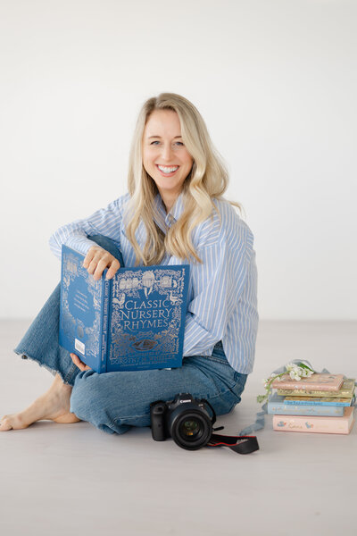 a woman sits on the floor with a white backdrop as she holds open a Children's Nursery Rhymes book. A photography camera and a stack of books and flowers sit next to her