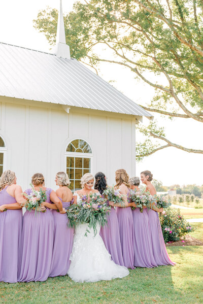 Bridesmaids in purple dresses with bride holding large bouquet of a lot of greenery and purple and white flowers taken by Greenville photographer