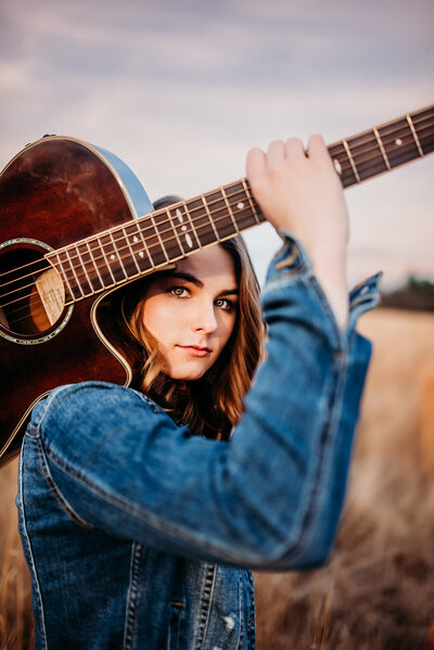 a senior girl, holding her guitar on her shoulder looking through the hole of her arm and guitar