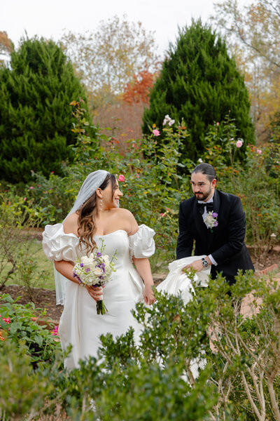 A groom holds the train of his bride's wedding dress as they walk through a garden of flowers and greenery in the suburbs of Raleigh NC.
