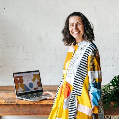 A woman with dark, shoulder-length hair stands next to a table with an open laptop. She is wearing a vibrant, patterned dress with a mix of colors including yellow, orange, and black. The background is a neutral, textured wall.