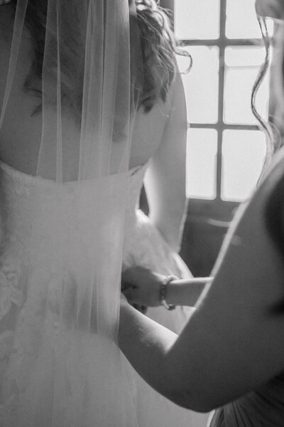 A black and white closeup image of bridesmaid helping bride zip her wedding dress in her grandmother’s home. Taken by Richmond Wedding Photographer, Caitlin Wilcox