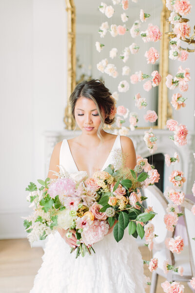 Bride holding a big pink bouquet with greenery standing next to strings of pink flowers hanging from the ceiling