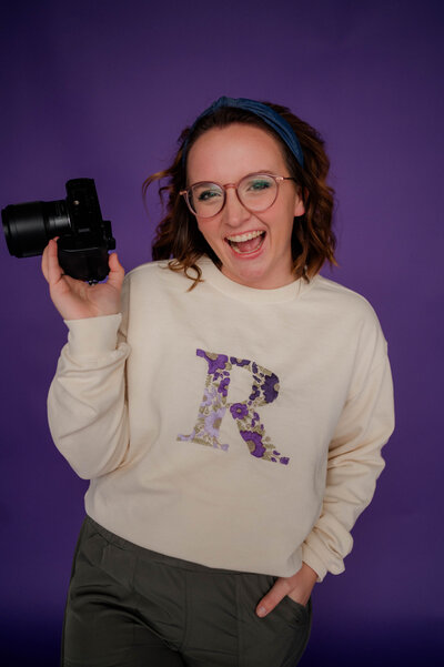 Rachel Hahn, owner, smiles with her camera in hand in front of a purple backdrop wearing a crewneck with the letter R in purple flowers.