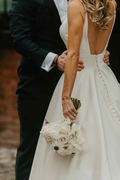 Groom holding the bride’s waist as she holds a bouquet of white roses, showcasing the back details of her elegant wedding gown.