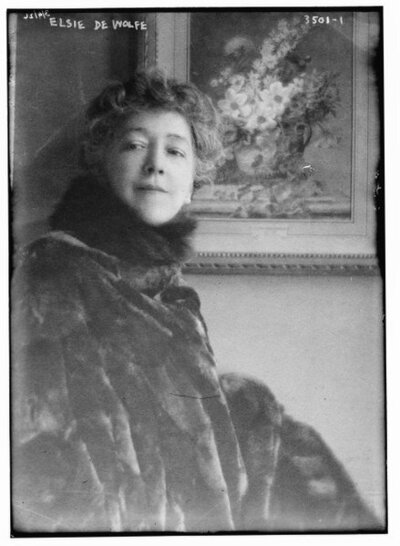 A black and white portrait of Elsie de Wolfe, the American interior decorator and socialite, draped in fur, against the backdrop of a framed floral painting, reflecting her era-defining elegance and impact on luxury interior design.