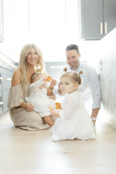 A Dallas family of 4 portrait of them sitting of their kitchen floor interacting with their two baby's that are snacking on some cookies as their little toddler looks at the photographer.