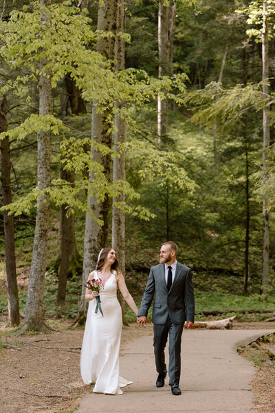 Couple walking while holding hands with green trees all around them.