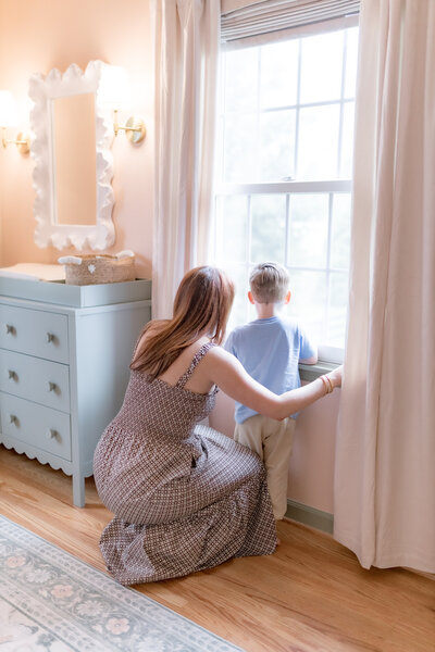 mom and little boy looking out the window in baby sisters nursery for lifestyle family session with natural light 