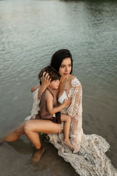 Mother and son embracing on a beach, showing affection, with water gently washing around them, warm tones