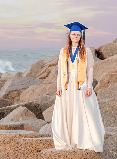 A Cape Fear Community College senior in graduation attire standing on rocks with the ocean as a backdrop in North Carolina. This elegant image captures the essence of destination graduation photography, ideal for high school seniors looking for memorable senior photos at the beach.