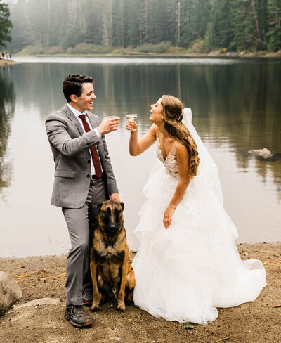 a bride and groom toasting champagne next to an alpine lake with their dog