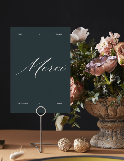 A dark card with the word "Merci" in script font is displayed on a holder next to a floral arrangement in a stone vase on a table—an elegant touch inspired by Sweet Pea Ink Creative’s brand strategy.
