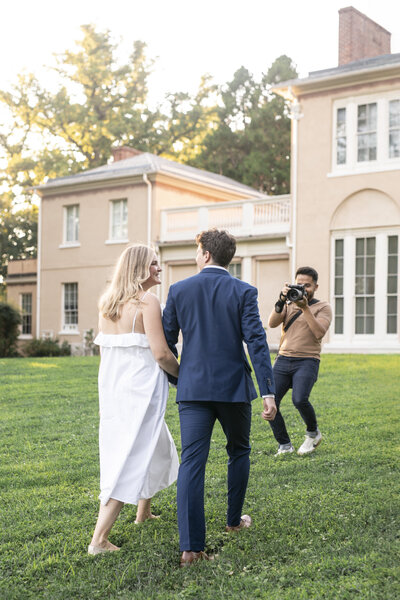 A couple walks hand in hand on a grassy lawn, approaching a photographer. The woman wears a white dress, and the man is in a blue suit. They are in front of a large beige house with tall windows and surrounded by trees.