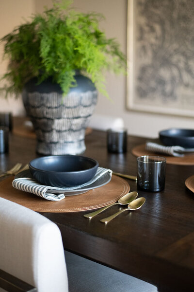 a place setting on a dining table with a navy bowl, brown charger, and gold utensils