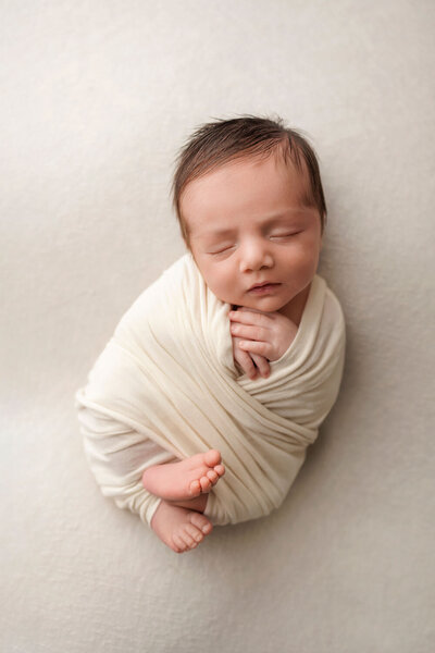 A newborn baby is peacefully sleeping, wrapped snugly in a creamy white swaddle. The baby is lying on a soft, neutral-colored background, with their tiny feet peeking out from the wrap. The baby’s hands are gently placed near their face, and their expression is serene and content.