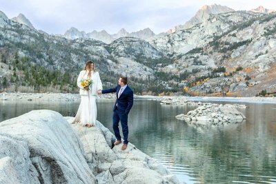 A groom holds his brides hand as he helps her walk down a rock in front of a beautiful lake view on their mammoth lakes wedding day. 