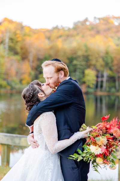 jewish couple kissing with fall floral bouquet and lake in background by asheville wedding photographer