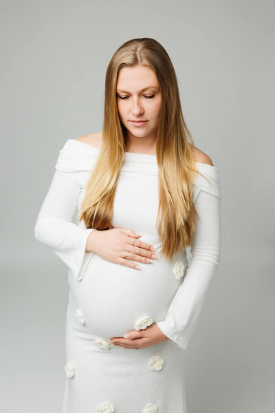 pregnant mother in a cream dress on a cream backdrop posed and smiling softly showing. off her pregnant belly in maternity studio session in harrisburg