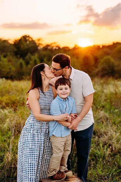 Family of three photographed during their Sunset Family session in a field in Memphis, TN by Jen Howell Photography. Mom and Dad kiss, and child smiles at the camera. The sky is pink and orage.