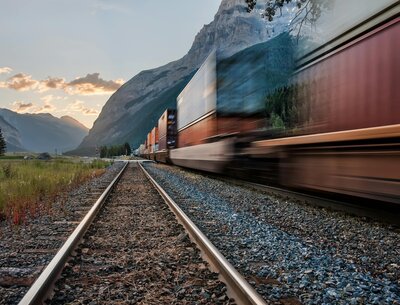A freight train speeds along tracks through a mountain landscape at sunset, representing Mya Andersen's goal of traveling all 50 states by train 
