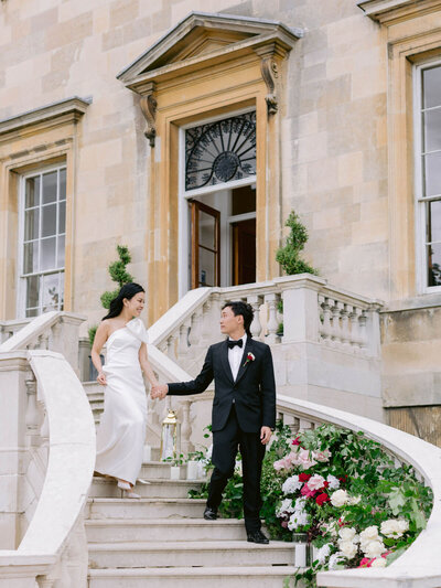bride and groom holding hands walking down the staircase decorated with luxury red and white roses at botleys mansion