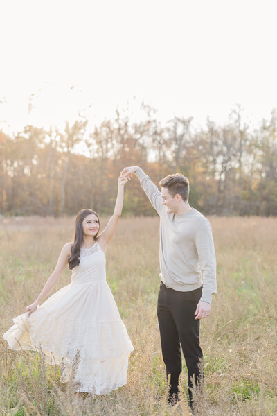 A newly engaged couple dances in a field of tall grass at sunset in a white dress and grey shirt