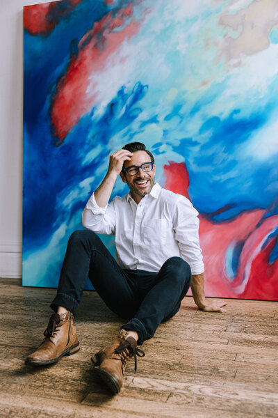 Branding photo of a man wearing glasses sitting in front of a painting for a small business in Pittsburgh, PA.