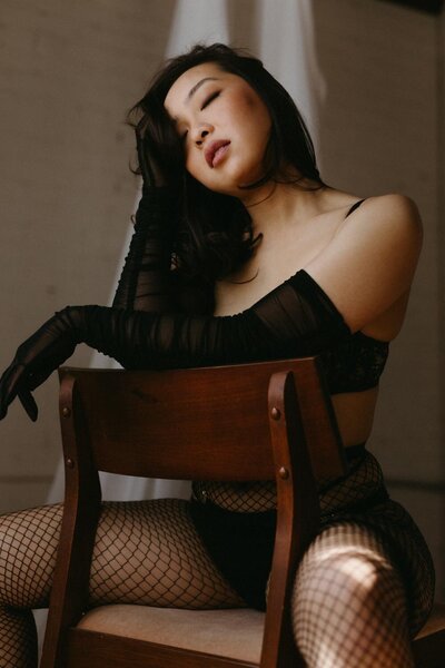 A woman sitting backwards in a chair with sheer gloves and fishnet stockings