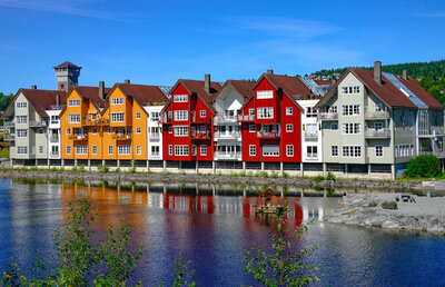 Row of colorful wooden houses along the riverside in Trondheim, Norway, reflected in the calm water under a clear blue sky.