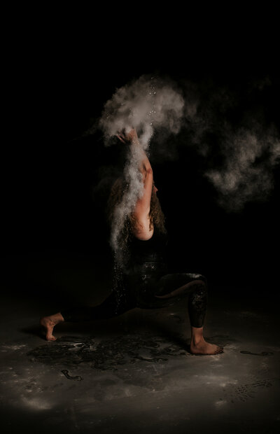 Alex Paige in a yoga pose in front of a black wall with chalk smoke around her