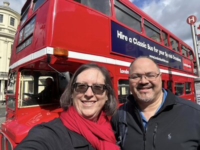 A man and a woman, wearing a red scarf, are standing in front of a vintage red London double-decker bus.