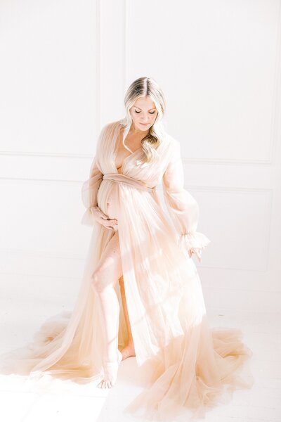 A Charlotte maternity photographer captures a pregnant mom  in a beige gown posing gracefully in front of a white background.