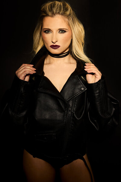 Pop star headshot of tina win. She's standing in front of a black wall, wearing black leather jacket and black jean shorts.