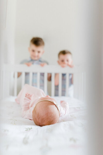 Two brothers peeking on their baby sister through the crib as she sleeps soundly during a lifestyle newborn session in Charlotte, North Carolina.