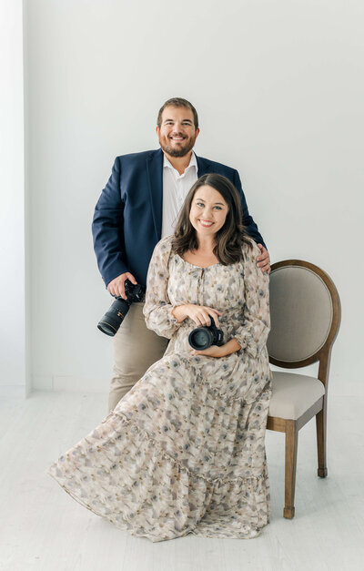 Katiera sitting on a chair holding a camera with hunter standing to her side holding a camera while they smile
