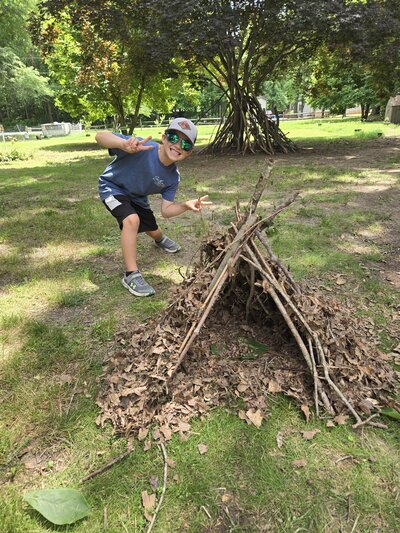 A child standing behind a teepee that they constructed with sticks and leaves