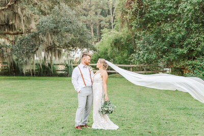 Newlyweds stand in a lawn smiling at each other as the veil blows in the wind behind the bride