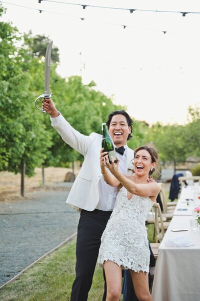 A bride and groom laughing together as they pop a champagne bottle at their California vineyard wedding.