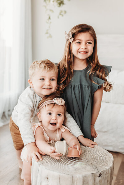 three siblings standing together at milestone photographer studio  leaning on tree stump prop