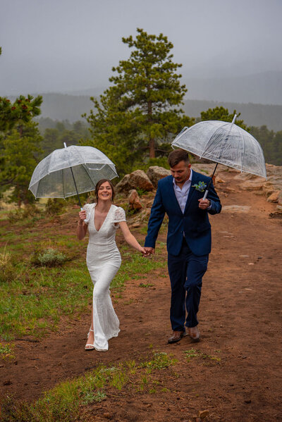 A bride and groom hold clear umbrellas and walk together with an overcast sky.