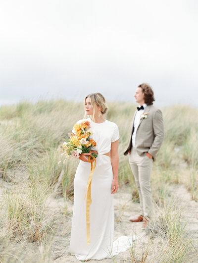 bride holding bouquet and the groom standing behind her both looking off into the distance at Cannon Beach