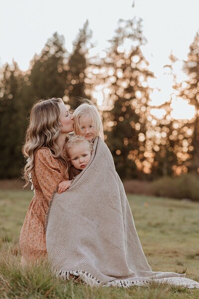 Two toddler sisters wrapped in a blanket in a meadow at sunset being kissed by mom