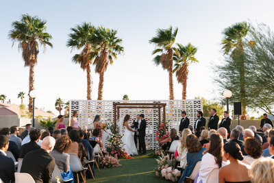 Outdoor wedding ceremony at the Lautner Compound in Palm Springs, California, sitting under palm trees, with guests seated around a floral-decorated altar in a colorful setting during golden hour.